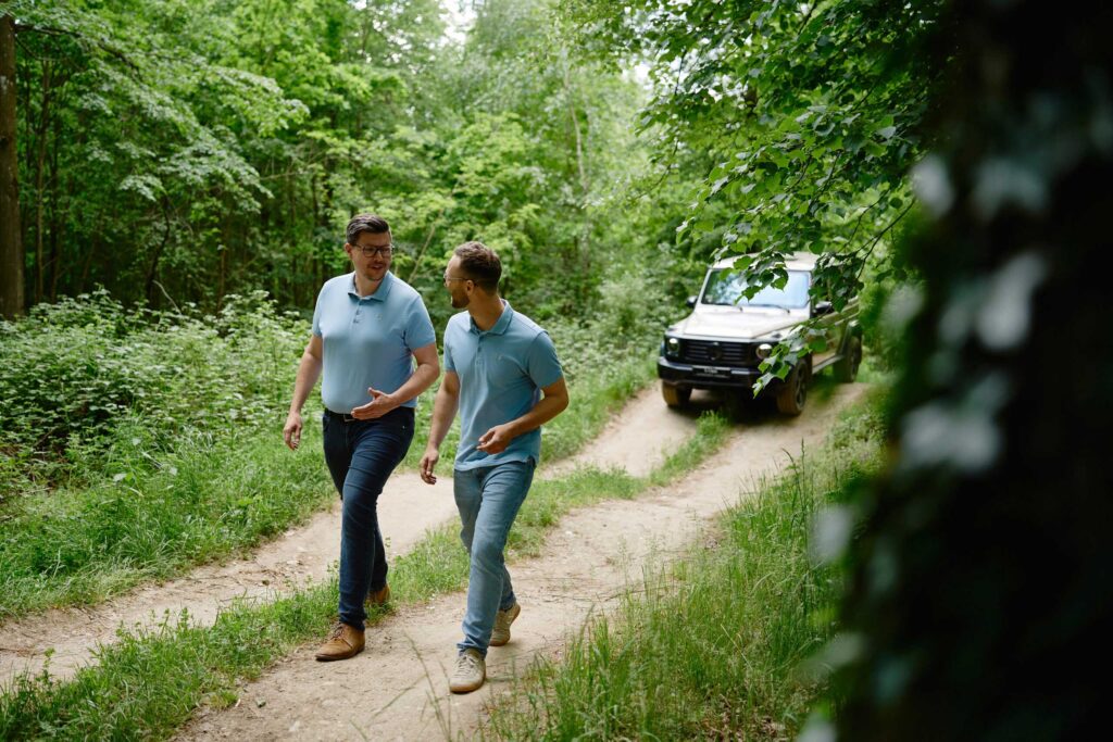 Zwei Personen gehen einen Forstweg entlang, im Hintergrund sieht man eine Mercedes-Benz-G-Klasse.