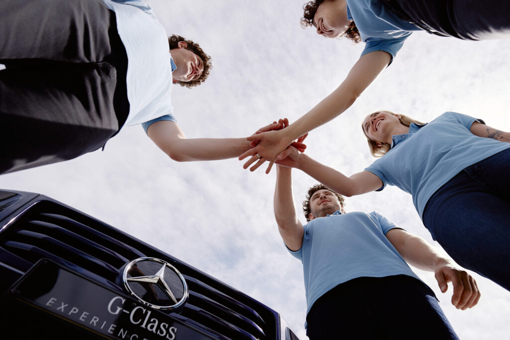 Mehrere Personen legen die Hände übereinander vor einer Mercedes-Benz G-Klasse.